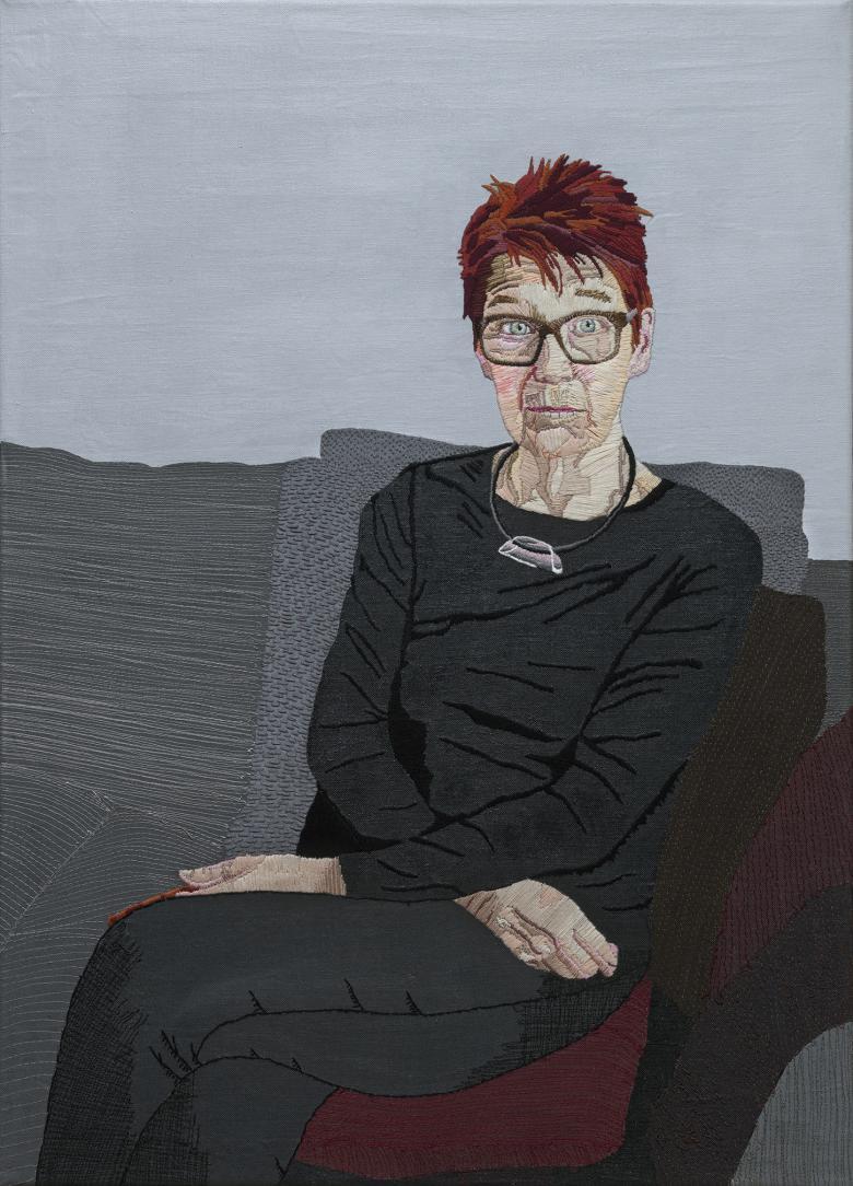 An embroidered portrait of a woman seated on a grey sofa, with her arms crossed in her lap.