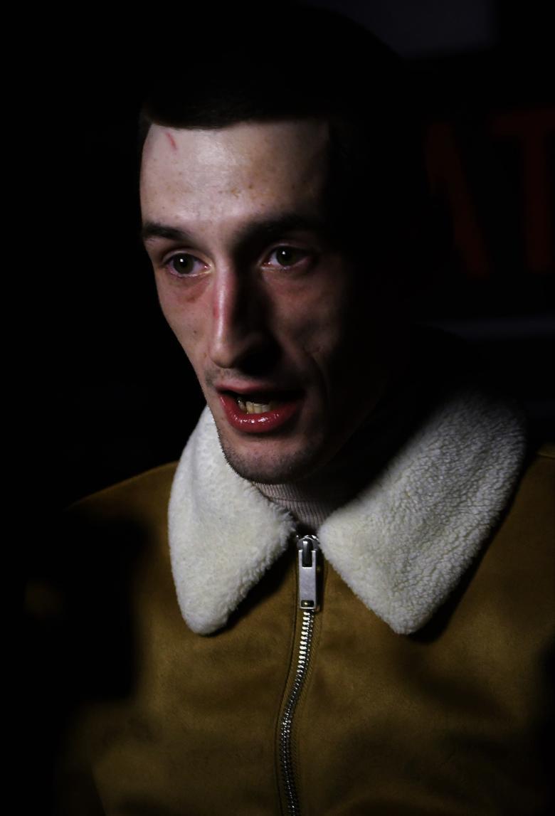 A face peers out of the shadows. The man wears a zipped up jacket with a white furry collar. His mouth is partially open, and his eyes look past the viewer, to the left of the frame.