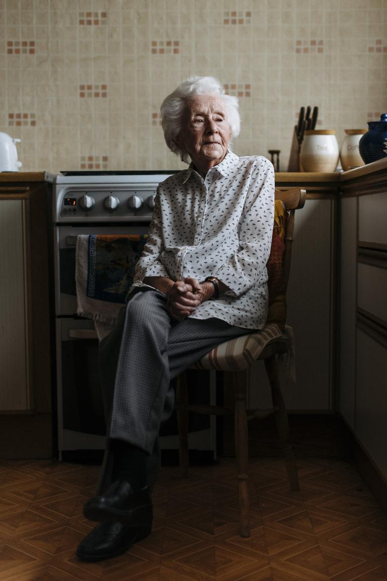 A woman sits on a chair in her kitchen, her hands clasped in her lap. Her legs are crossed, and she looks down, away from the viewer.  