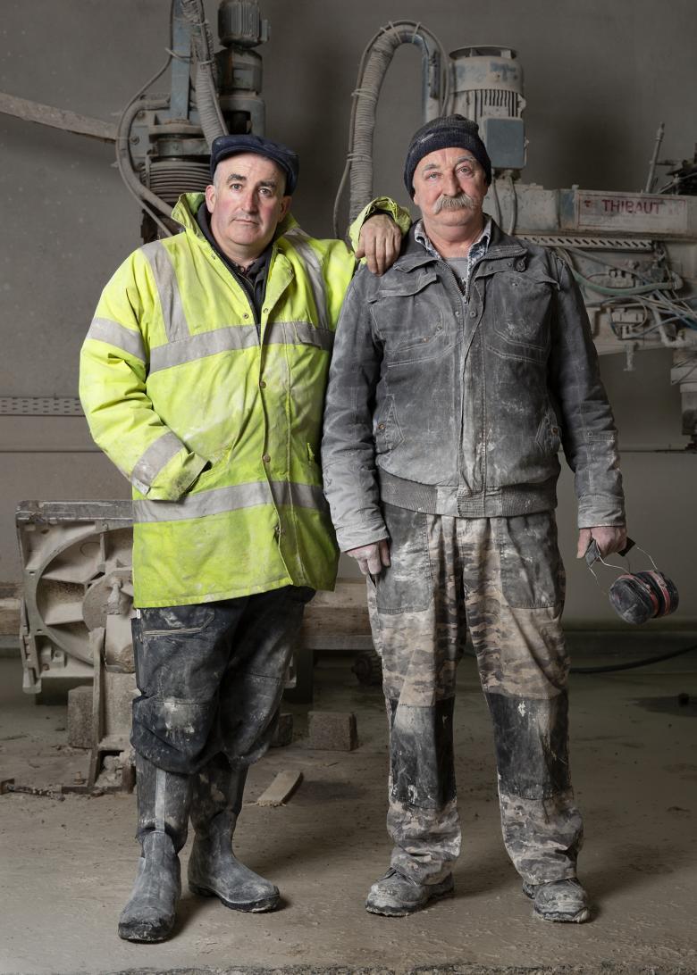 Two men stand in front of industrial-looking equipment. They wear work clothes and boots, and are covered in dust. The man on the left wears a high-vis jacket and rests his hand on the shoulder of the second man.