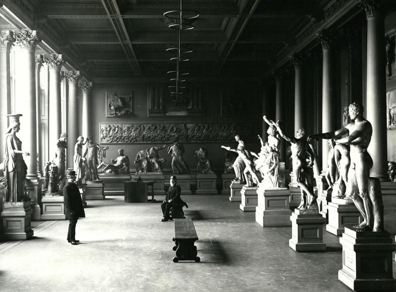 A black and white photograph of a room filled with plaster casts of sculptures. There are two me in the room - one sits on a bench in the middle of the room, the other stands on the left, facing the seated man.