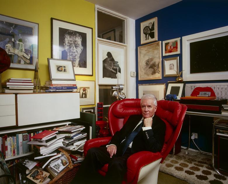 Photo of Alexander Walker in his apartment sitting in a red leather armchair with lots of art hanging on the walls