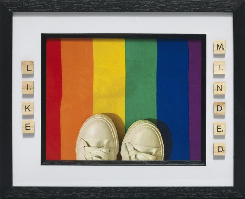 Birds-eye view of white sneakers standing on a rainbow-coloured rectangle. The words like minded are spelled out in Scrabble tiles.