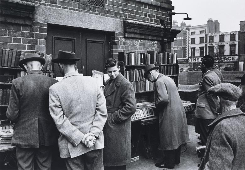 A group of men, most wearing hats, some with the hands behind their backs, peruse a bookstall in a city.