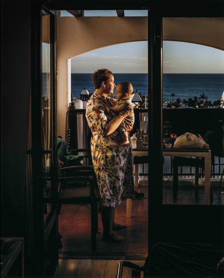 Portrait photograph of a woman wearing a robe and holding baby, standing in a room with a large window with a view of the sea