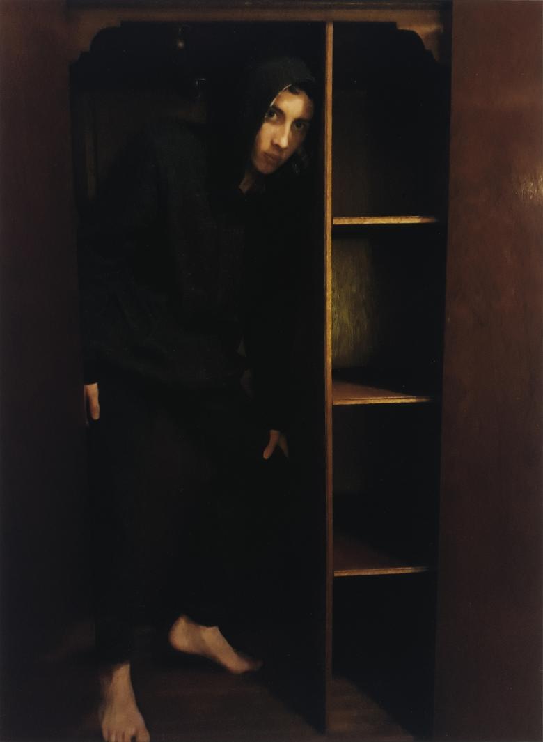 Portrait of a young man wearing black trousers and black top with hood up standing in the shadows in an empty wardrobe