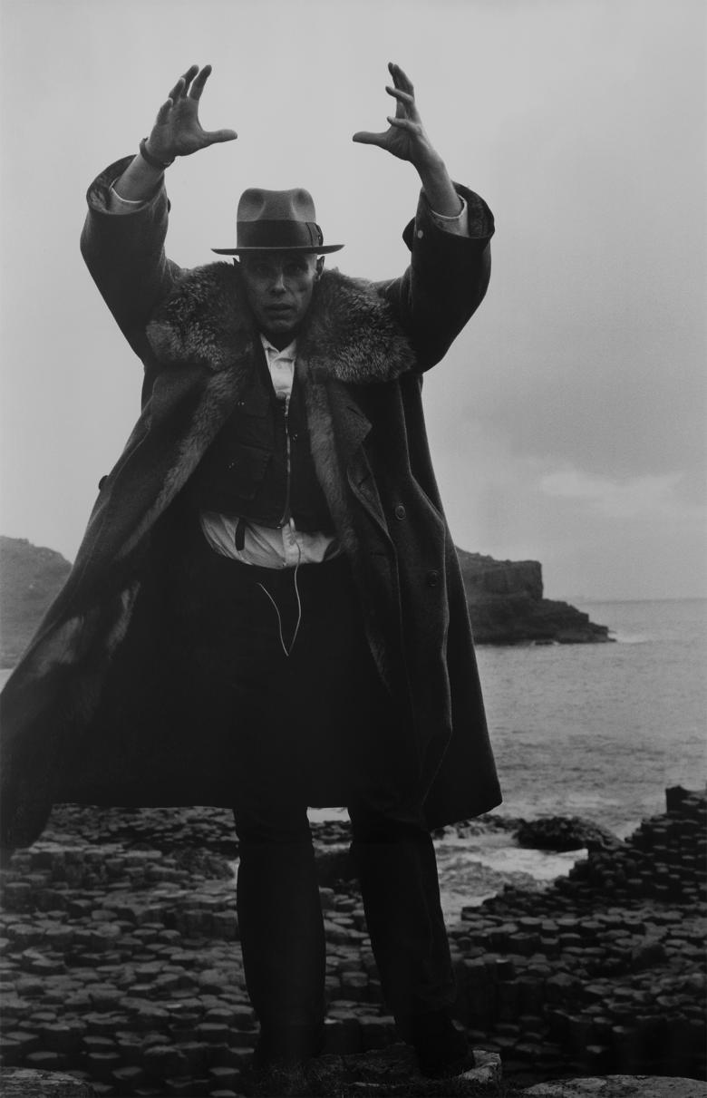 Joseph Beuys stands in front of a windswept landscape, his hands raised above his head, wearing a long coat and a hat