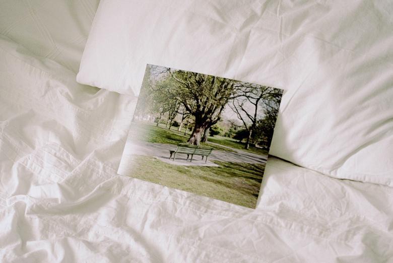Photograph of a photograph lying on an unmade bed with white sheets. The photo is of a man sitting on a park bench.