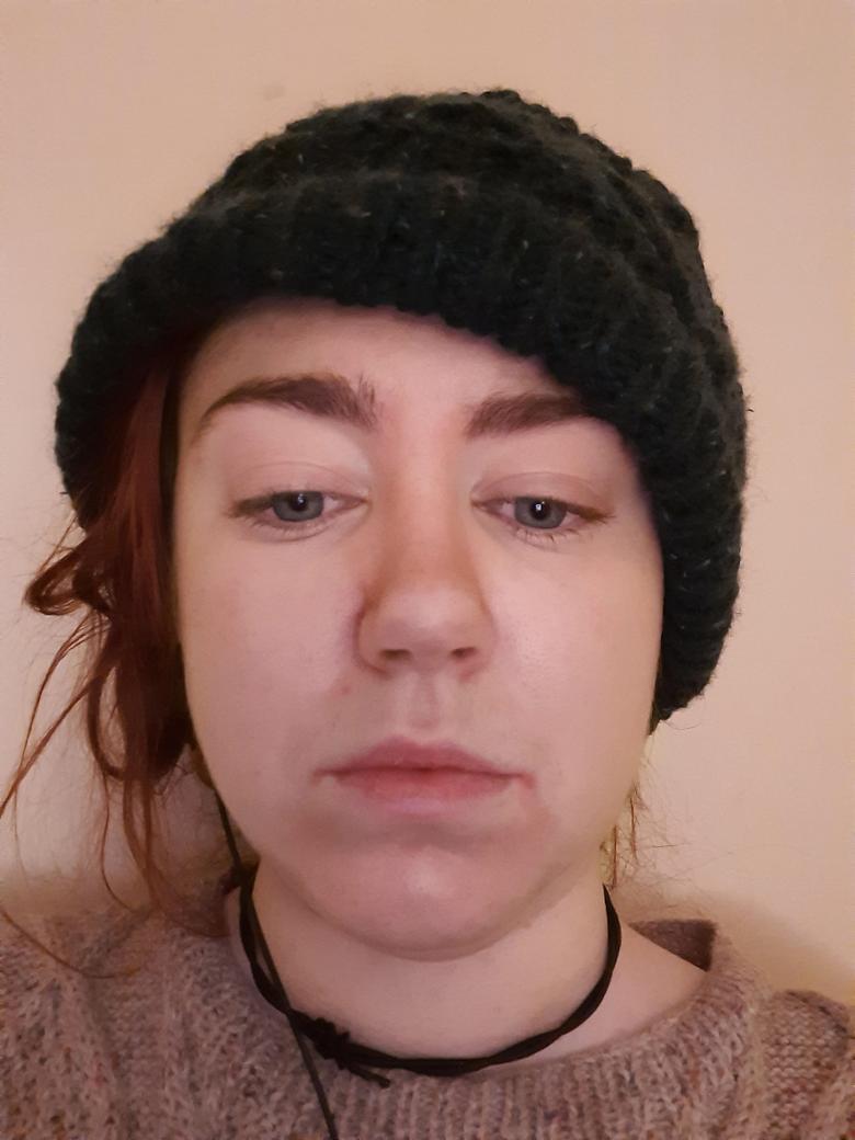 Video still, close-up view of a woman's face. She is wearing a black woolen hat and her eyes are cast downwards.