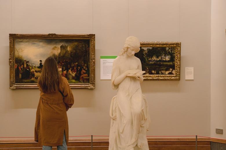A woman looks at a landscape painting, while standing next to a marble sculpture of a young woman reading. 