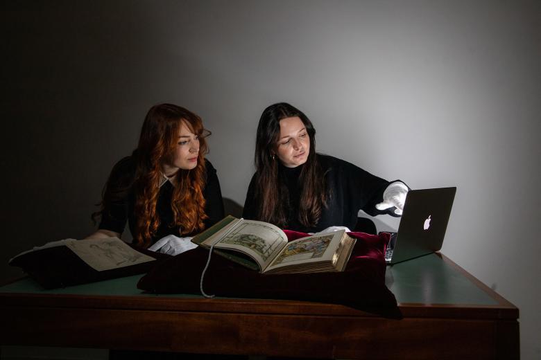 Two researchers looking at a laptop with rare books lying on the table on cushions
