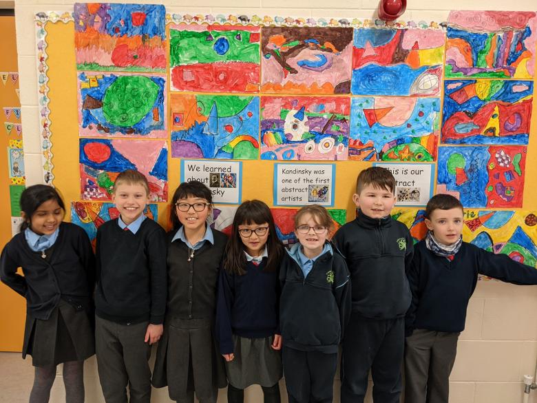Seven children in a classroom standings in front of a dozen artworks done by pupils. 