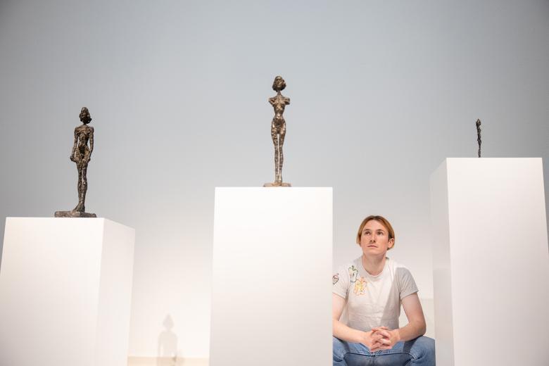 Richard Malone posing next to three white plinths displaying three sculptures by Alberto Giacometti