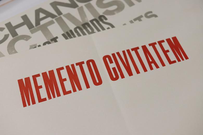 A sheet of paper with memento civitatem printed in red block capitals, with another sheet of paper visible beneath with fragments of words including activism and change