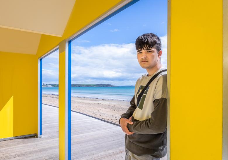 Photograph of a male figure with dark hair and a dark green jumper leaning against a yellow structure with his back to the beach