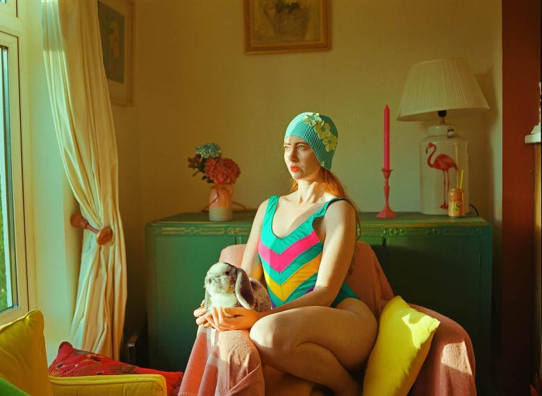 Photograph of a female figure with red hair wearing a teal striped swimsuit and hat sitting in a pink chair holding a toy rabbit