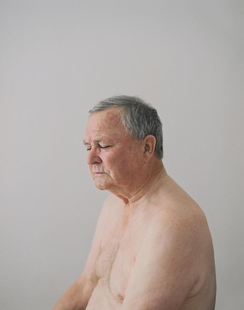 Photograph of an older man's head and shoulders with grey hair and his eyes closed sitting nude on a grey background