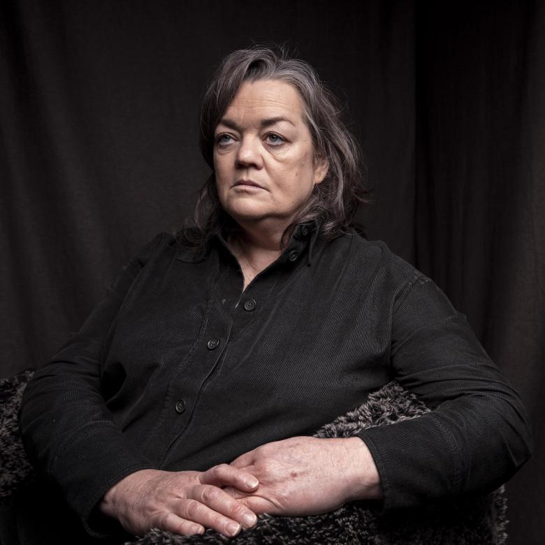 Photograph of a female figure with short grey hair and a black top on a black background