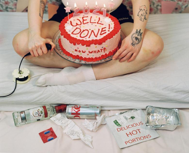 Photograph of a figure with tattoos sitting cross-legged on a bed holding a cake with various rubbish nearby