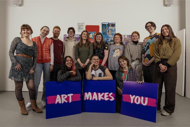 Photograph of a mixed group of young people, with three purple signs with the statement 'Art Makes You'