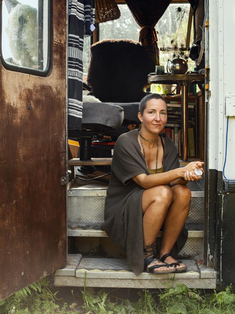 A photograph of a woman sitting on the steps of a caravan. She is wearing a long grey cardigan and has bare legs. Her hands are clasped in front of her.