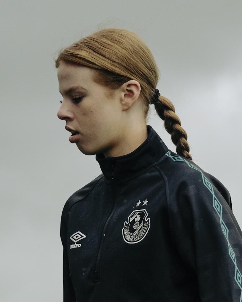 A young woman in a crested tracksuit top stands in profile, eyes cast down. Her red hair is tied back in a long plait.