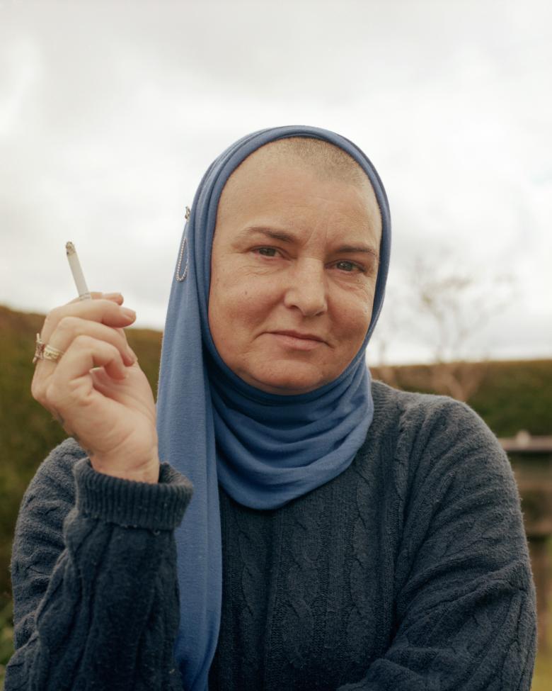 A photographic portrait of a a woman. She wears a dark blue knitted jumper and a blue headscarf. She holds a cigarette aloft in one hands. Behind, a grey sky and some green fields. 