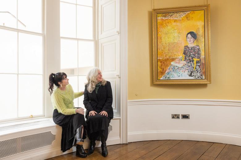Photograph of two women, one wearing black, the other wearing a green top, sitting in a yellow room looking at a painting