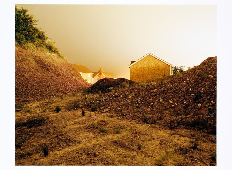 A photograph with a golden hue, showing a landscape with fresh earth overturned and some houses in the distance