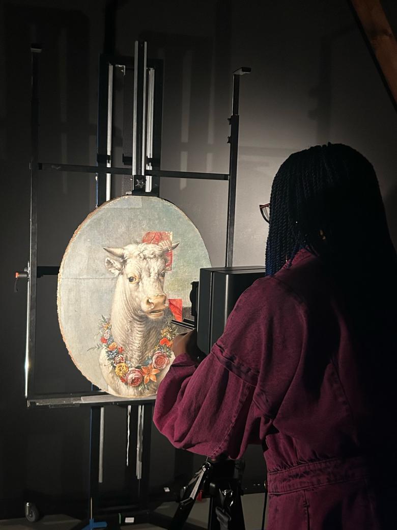 A small oval painting of a bull stands on an easel; someone stands in front of it taking an infrared reading