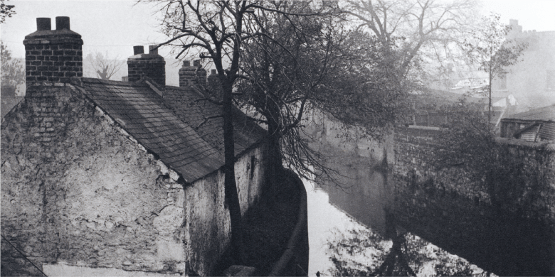 A black and white photograph of cottages beside a river. Bare trees beside the cottages are reflected in the river.