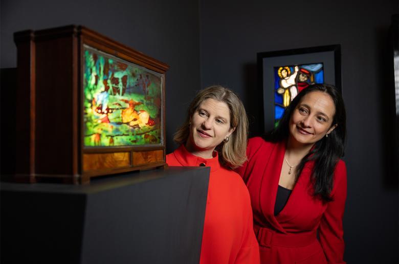 Photograph of two women, both wearing red dresses, looking around a corner at a piece of decorative stained glass.