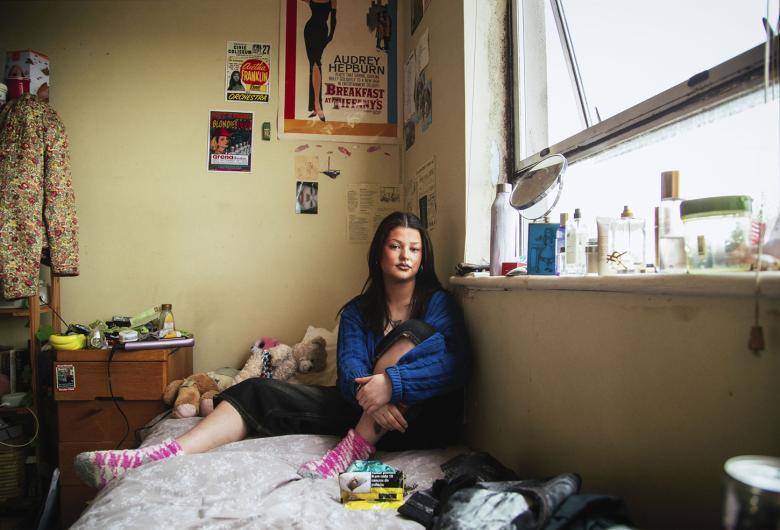 A photograph of a young woman sitting in her bedroom, surrounded by her belongings. On the wall, a number of posters and cuttings hang including a Breakfast at Tiffany's poster; on the bed itself are a few stuffed animals, and various cosmetics line the window sill.