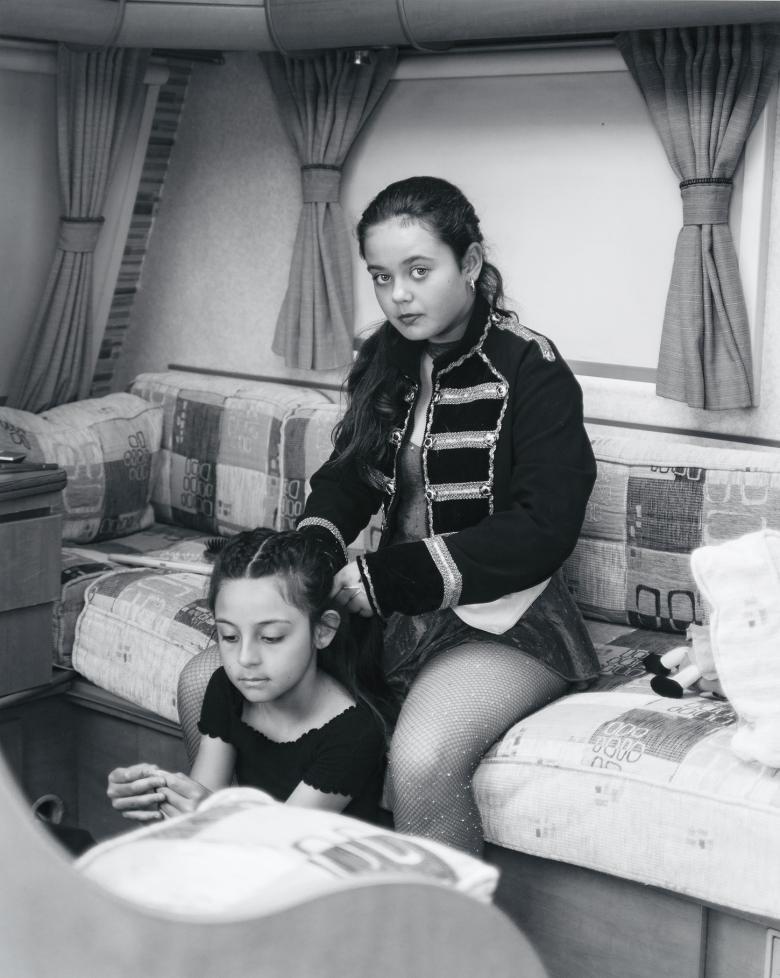 Sitting on a sofa in what appears to be a mobile home, a young woman with long dark hair, wearing a ringmaster's jacket, sits plaiting a younger girl's hair. She looks directly at the camera. 