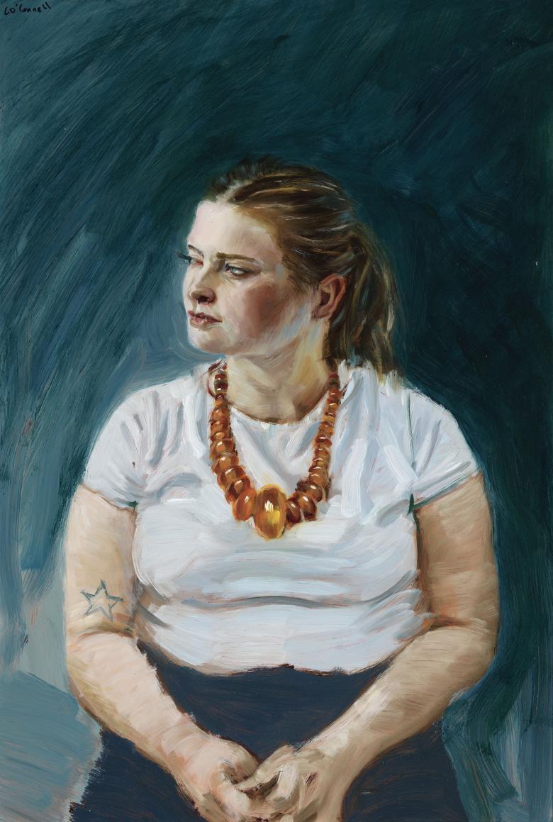 A woman sits against a dark blue background, her hands clasped in her lap and her head turned slightly to the left. She wears large amber beads around her neck, and a tattoo of a star is visible on her upper arm.
