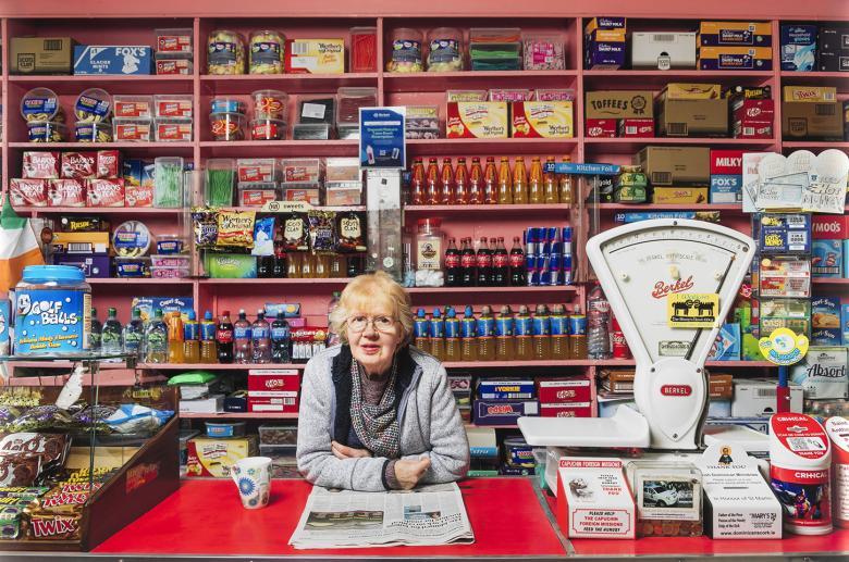 A woman stands behind the red counter of a shop, her elbows on the newspaper open in front of her. Behind her, neatly stacked shelves of groceries, and on the counter a scales and more products.  