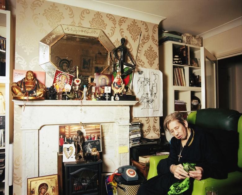 Shane MacGowan sits on a green armchair beside a fireplace. The mantle is filled with objects including a buddha, a picture of Jesus and other religious figures. A statue of Phil Lynott stands on the gas fire. 