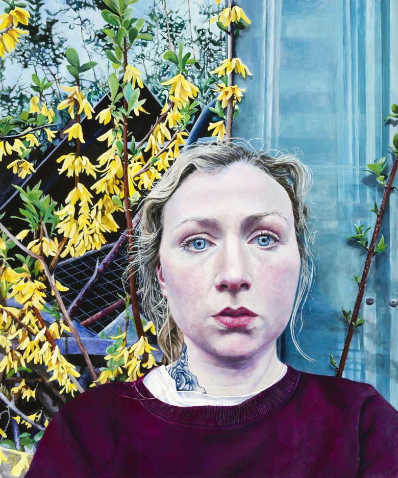 A woman with fair hair looks directly at the viewer. She wears a dark red top, with a white t-shirt underneath. A tattoo can be seen on her neck. Behind her the metal of a garden shed, and a bright yellow plant in bloom.