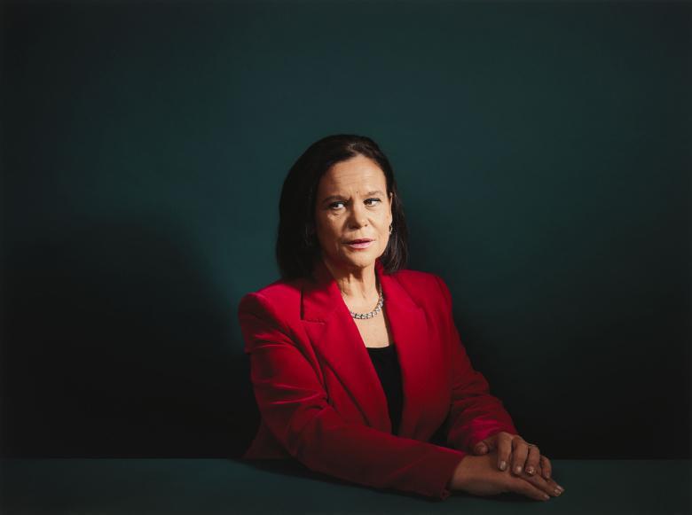 Dressed in a red jacket and seated at at table with her hands clasped in front of her, Mary Lou McDonald sits in front of a dark green wall.