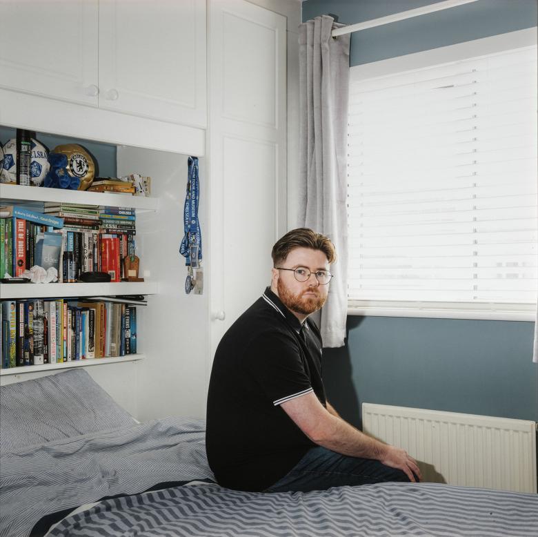 A young man sits on the bed in a neat bedroom, looking over his shoulder at the viewer. The shelf over the bed is filled with books and other belongings. 