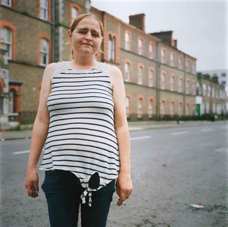 A woman in a sleeveless striped top and jeans stands slightly left of frame, looking directly at the viewer. In her hand she has a half-smoked cigarette. Behind her, a brown and red brick building, the site of a Magdalene Laundry.
