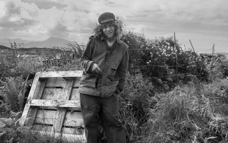A black and white photograph of a man standing in a windswept hilly landscape, with a wooden palette directly behind him. He has long curly hair underneath a cap, and is laughing at the camera.  