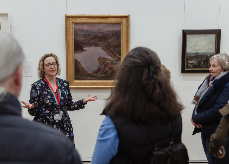 A guide stands in front of a painting, talking to a group of people who are looking at her. 