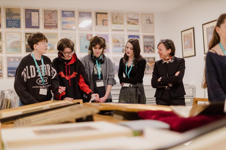 A group of young people standing around discussing prints and painting on a table.