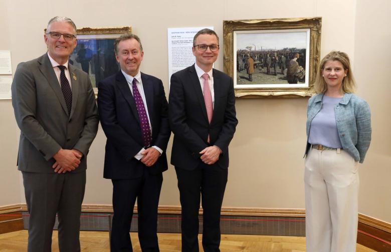 Three men wearing dark suits and a woman wearing white trousers and a blue top stand in front of a painting.