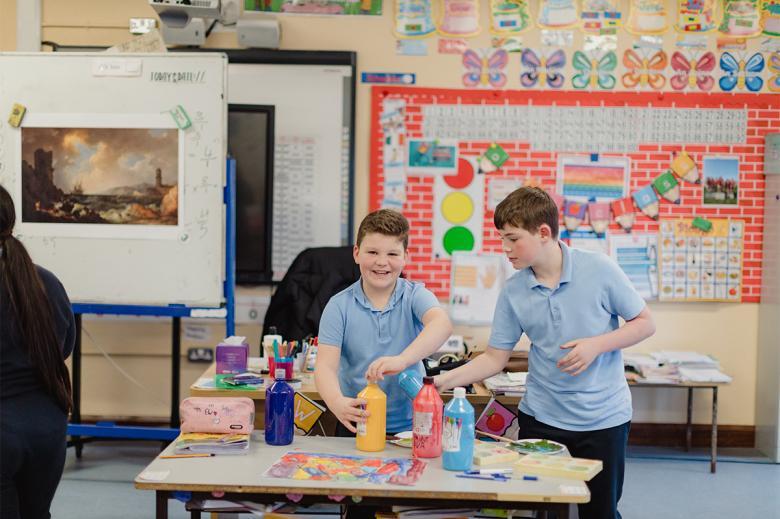 Two boys in blue polo shirts stand behind a table covered in art supplies, smiling at the camera