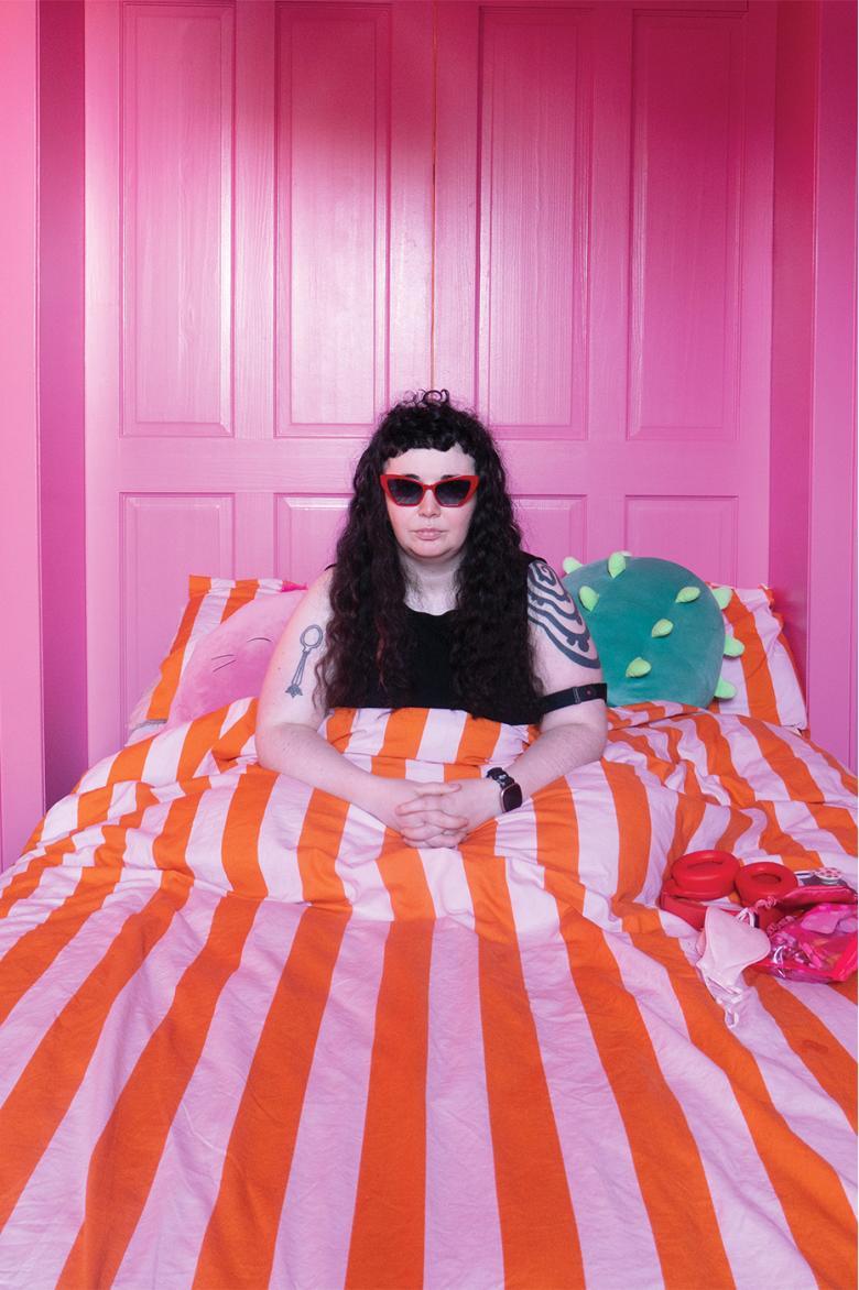A young girl with long black hair, sits up in a bed with a pink and orange striped duvet, in a pink room.