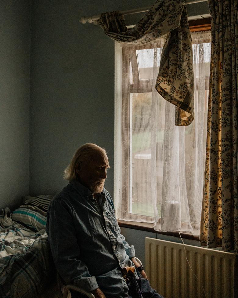 Photograph of an older man with a white beard, sitting in a small room in blue pyjamas