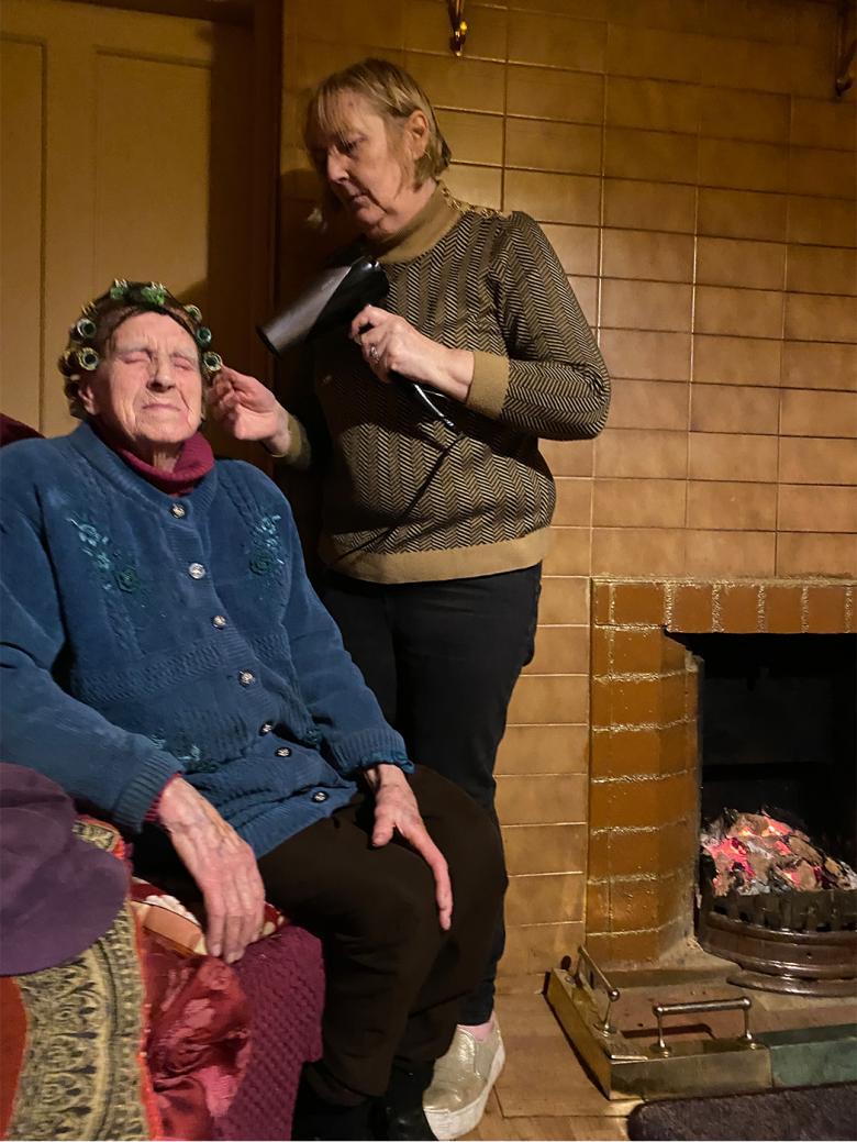 A photograph of a woman drying an older woman's hair in front of a fireplace