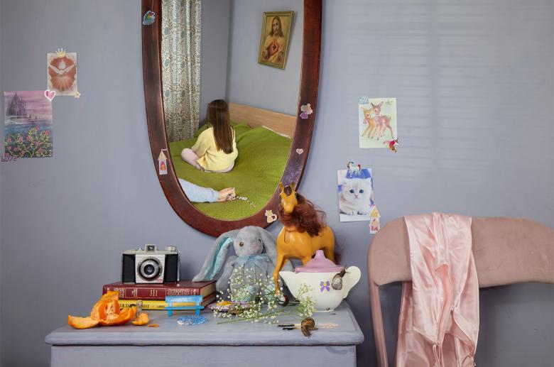 Photograph of a young girl with her back to us reflected in a mirror in front of an outstretched arm holding rosary beads. 